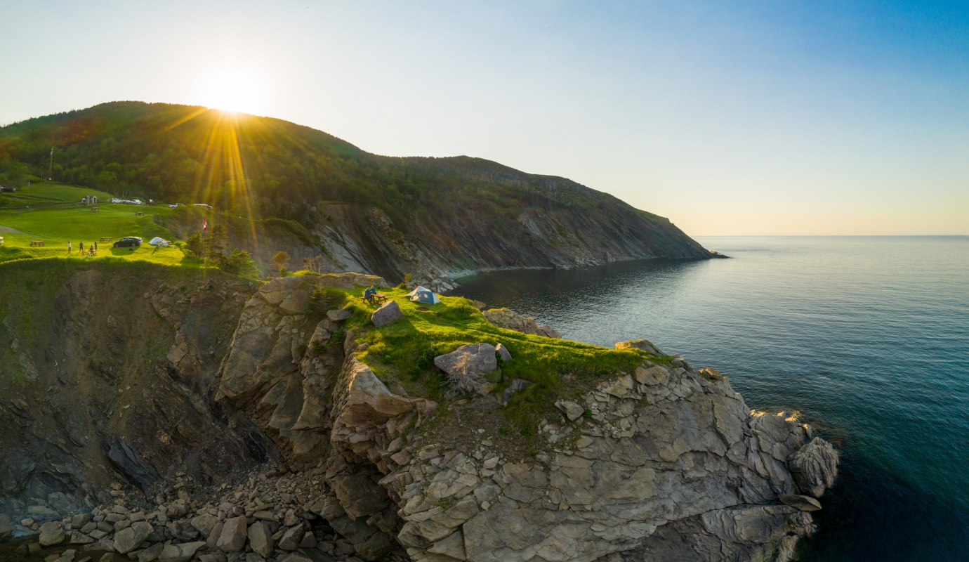sun peeks over mountain ridge as people camp by the ocean